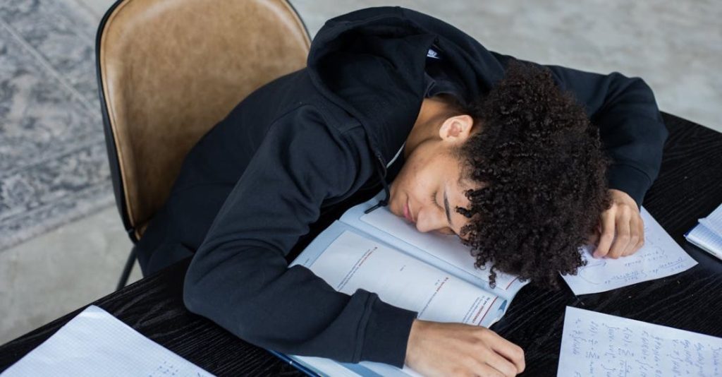 Task Overload - High angle of exhausted African American student resting on opened textbook and papers while preparing for exam