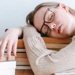 Task Lighting - Exhausted female student in eyeglasses and casual sweater sitting at table and sleeping on books on light room