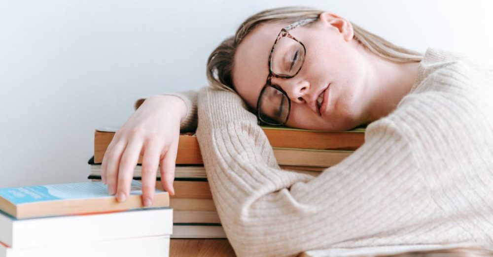 Task Lighting - Exhausted female student in eyeglasses and casual sweater sitting at table and sleeping on books on light room