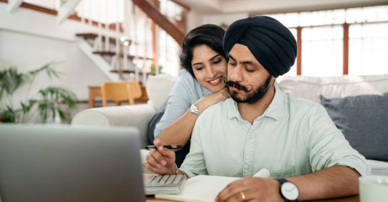 Remote Work - Cheerful young Indian woman cuddling and supporting serious husband working at home with laptop and counting on calculator